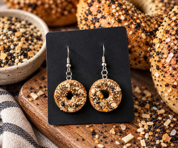 Bagel-style earrings on a card beside bagels and seeds