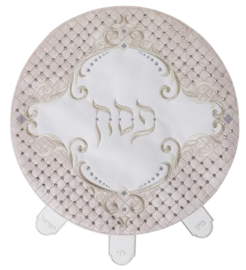 Round matzah cover with a blush quilted faux-leather border set with crystals, white velvet center embroidered Pesach in silver scrollwork and three labeled pocket tabs at the lower edge