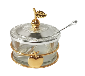 Round clear glass honey dish with a silver-tone embossed lid topped by a gold apple-and-leaf finial, gold band with an apple on the front and a silver spoon resting in the lid