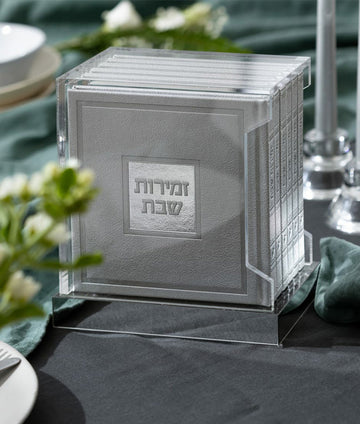 Simchonim Lucite stand holding hardcover zemiros in silver, shown on a table setting