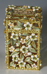 Gold-tone jeweled tzedakah box covered in white flowers with green leaves and small stones