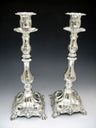 Pair of tall ornate silver plated candlesticks with scroll relief and square footed bases on grey