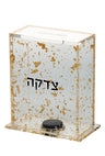 Lucite tzedakah box filled with gold flakes and black Hebrew Tzedakah on the front