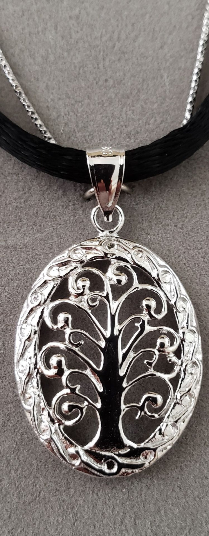 Oval Balinese-style Tree of Life pendant with dark backing, worn on black silk cord beside a silver chain