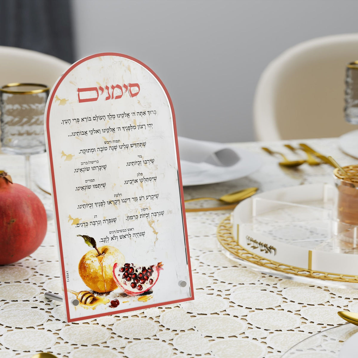 Simanim card standing on a lace tablecloth next to a pomegranate and gold-rimmed glassware