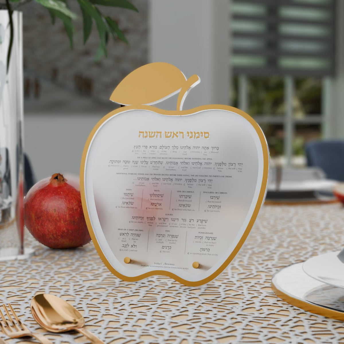 Gold simanim card on a patterned tablecloth beside a pomegranate and gold flatware