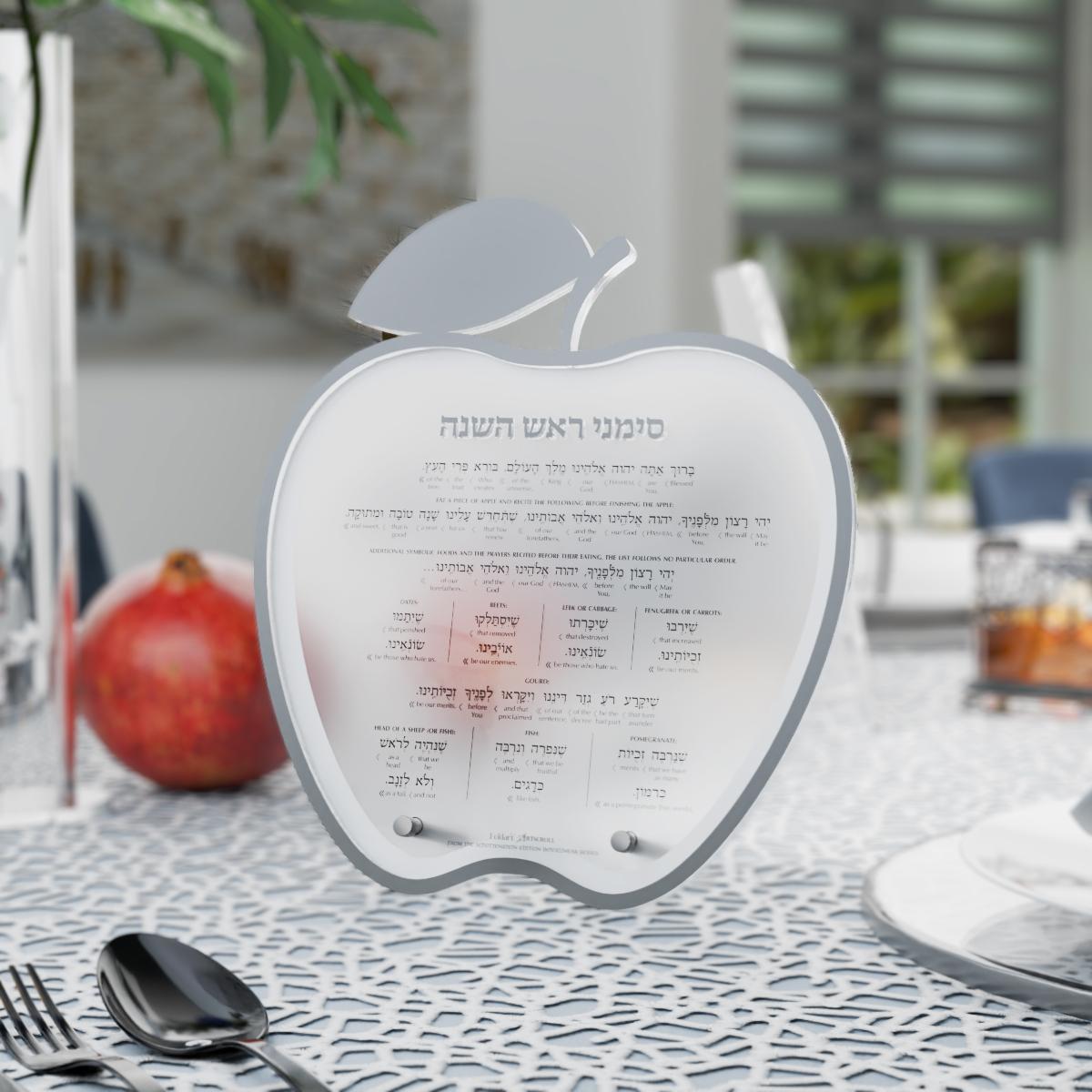 Silver simanim card standing on a set table next to a pomegranate and dark flatware