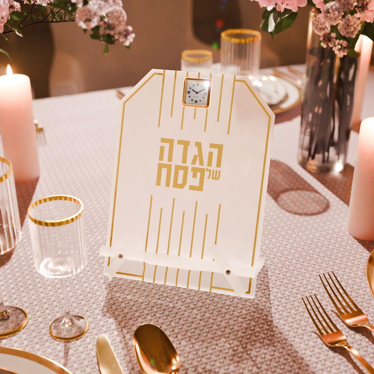 Gold Haggadah stand on a Seder table with gold-rimmed glasses, candles and pink flowers