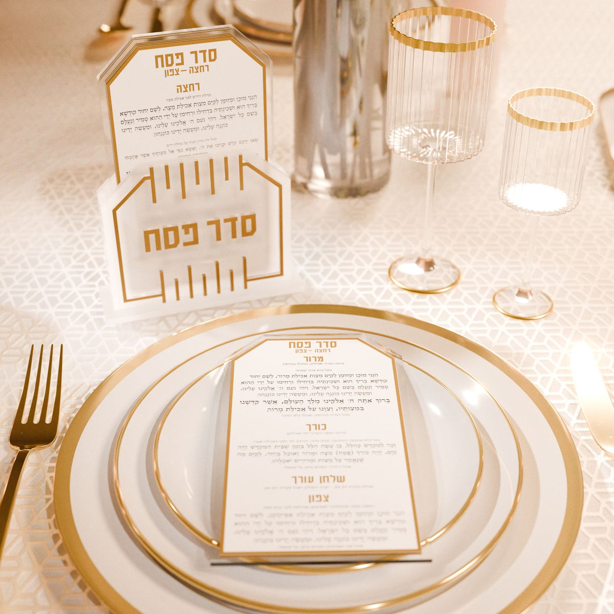 Gold Seder card on a place setting with gold-rimmed plates, holder and glasses behind it