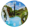 Round tempered glass wall clock printed with a waterfall pouring into a turquoise river between green forested hills, white numerals 1 to 12, black hands and white Hebrew lettering across the water