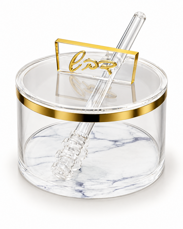 Round clear acrylic honey dish with a gold band at the rim and a white marbled base, lid closed with a raised gold cursive Hebrew handle and a clear acrylic dipper resting across it