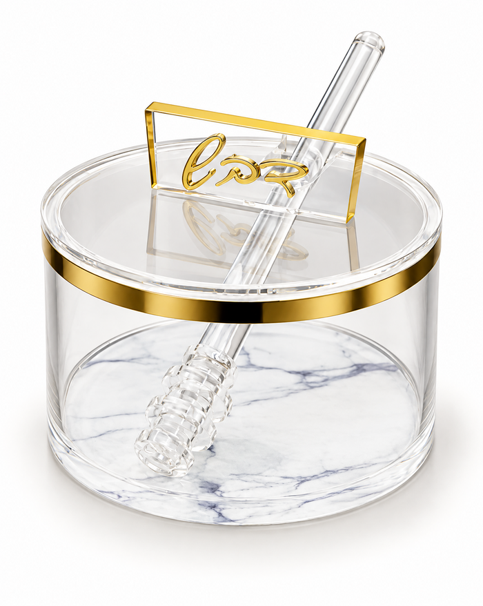 Round clear acrylic honey dish with a gold band at the rim and a white marbled base, lid closed with a raised gold cursive Hebrew handle and a clear acrylic dipper resting across it