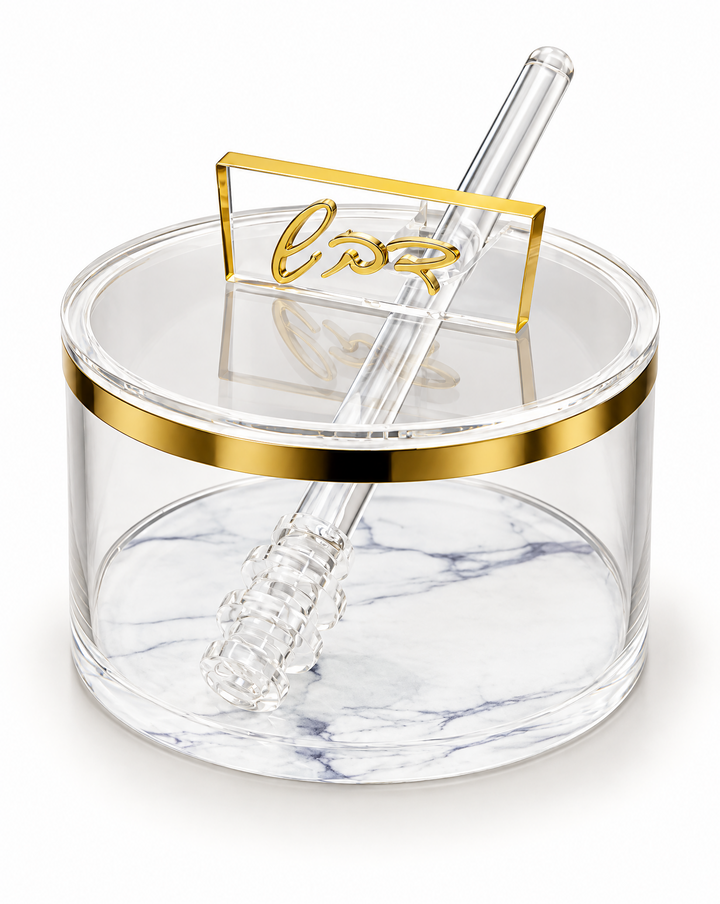 Round clear acrylic honey dish with a gold band at the rim and a white marbled base, lid closed with a raised gold cursive Hebrew handle and a clear acrylic dipper resting across it