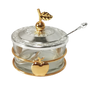 Round clear glass honey dish with a silver-tone embossed lid topped by a gold apple-and-leaf finial, gold band with an apple on the front and a silver spoon resting in the lid