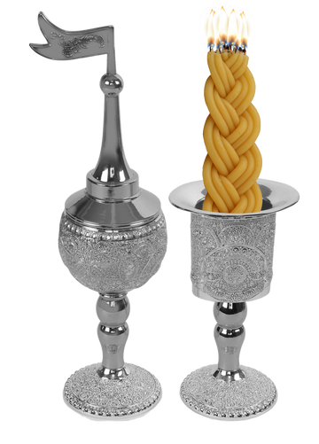Silver-plated filigree besamim holder with a pointed dome and flag finial beside a matching filigree candle holder with a lit braided havdalah candle in it