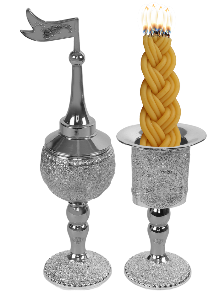 Silver-plated filigree besamim holder with a pointed dome and flag finial beside a matching filigree candle holder with a lit braided havdalah candle in it