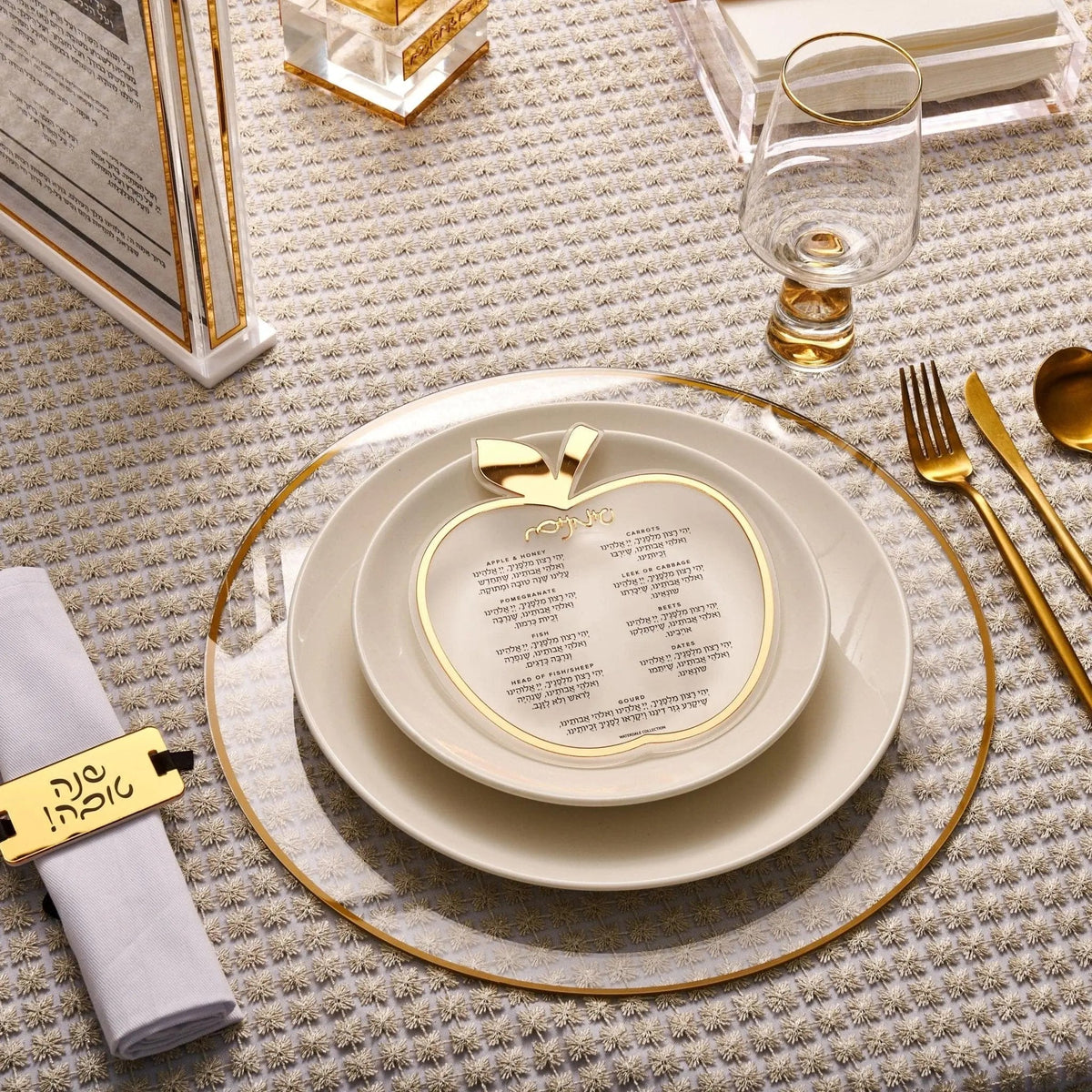 Apple simanim card standing on a Rosh Hashanah place setting with gold-rimmed plates and a Shana Tova napkin ring