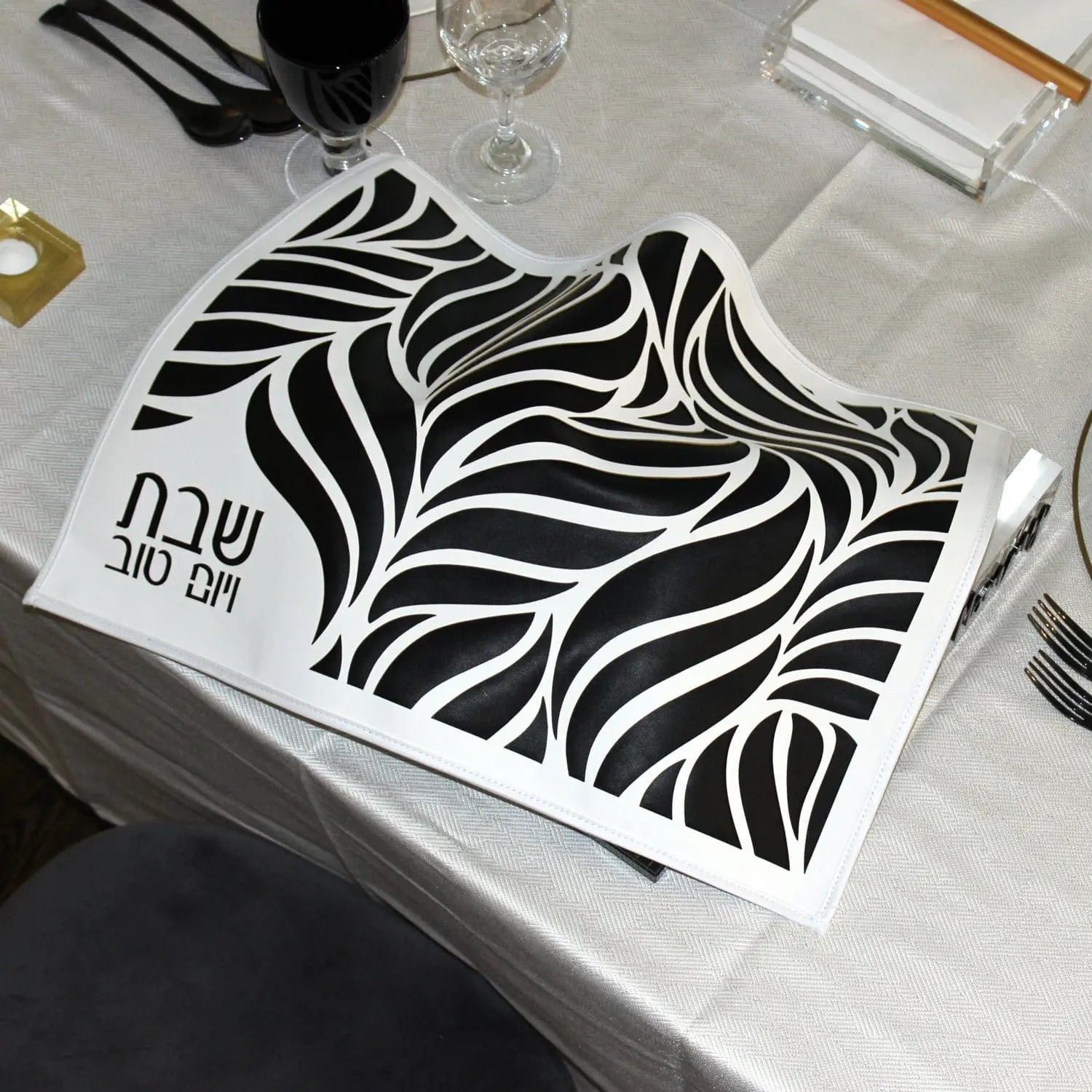 Black leaf challah cover draped over the challahs beside a black kiddush cup and wine glass