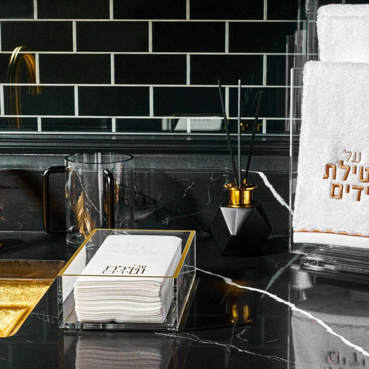 Black-handled lucite washing cup at a black-tiled kitchen sink with a lucite guest towel tray and hand towel